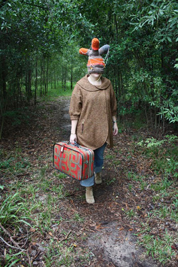 woman walking through the woods carrying a suitcase and wearing a knit hat the covers her face