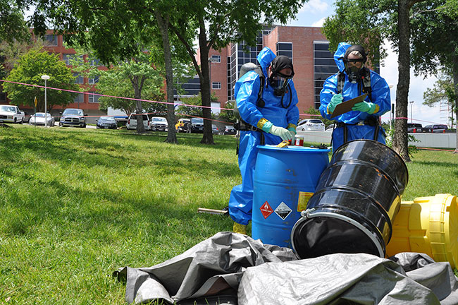 two men wearing blue hazmat suits standing next to several large barrels for chemical waste