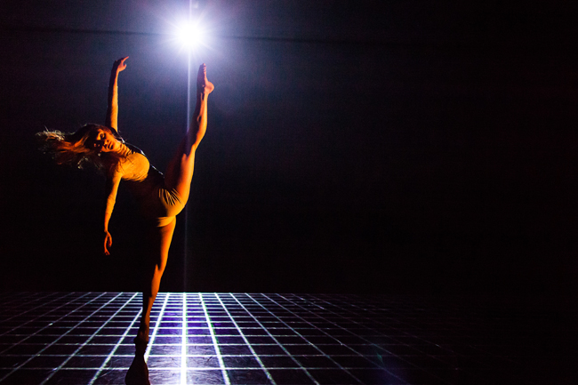 Dancer with leg high in the air bathed in a soft yellow light with a bright light above