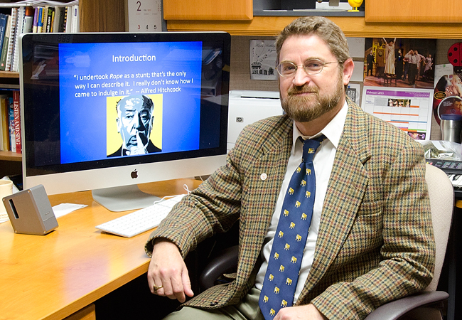 Clifton posing for a picture at his desk with a picture of Hitchcock on his computer